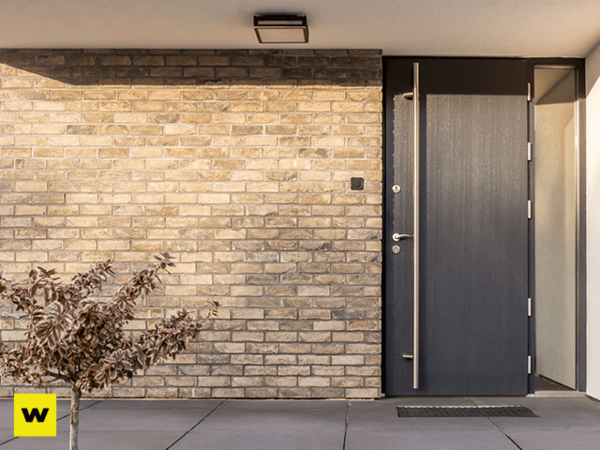 Moderne Haustür aus dunklem Metall neben heller Klinkerwand, Pflanze im Eingangsbereich.